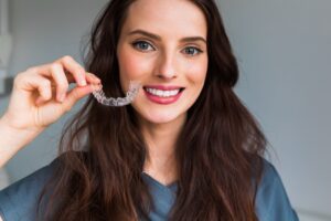 Woman with brown hair in blue shirt smiling holding Invisalign tray